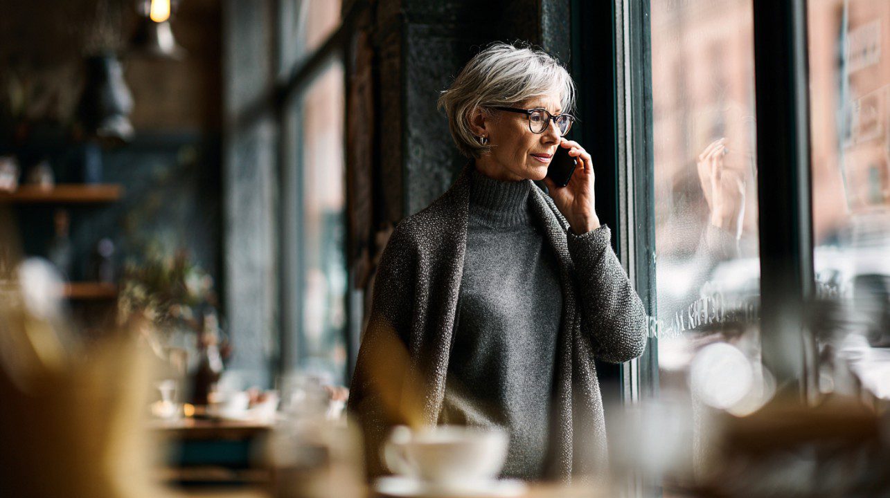 Mature woman with gray hair and glasses talking on her mobile phone while looking out the window of a cozy café.