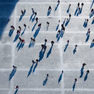 Top-down view of people walking on a cobblestone square, casting long shadows in bright sunlight.