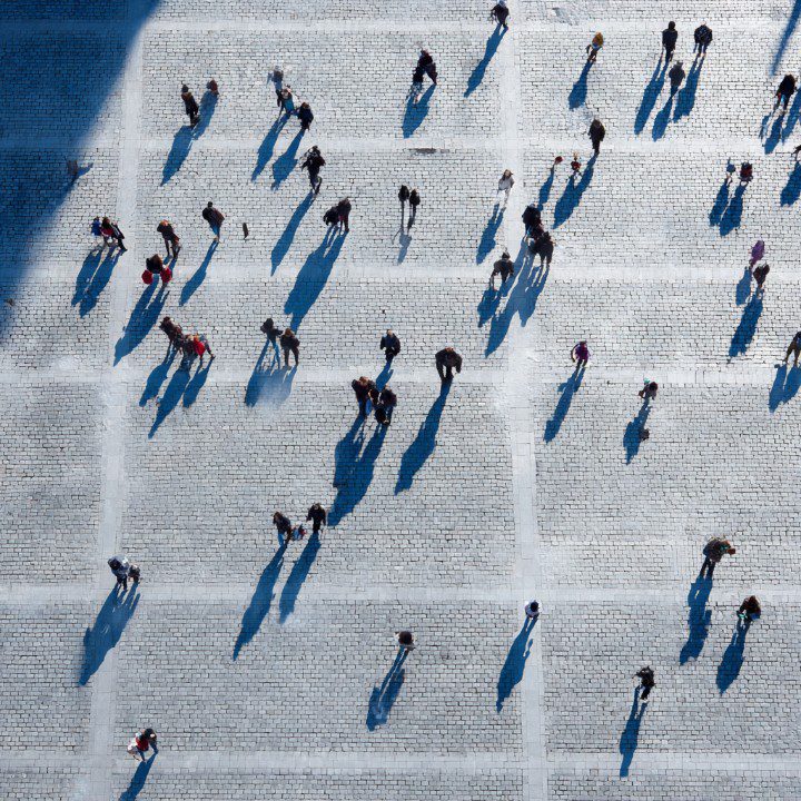 Top-down view of people walking on a cobblestone square, casting long shadows in bright sunlight.