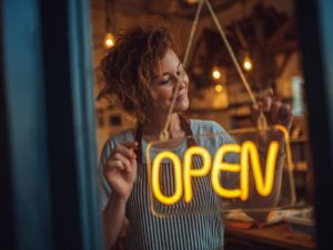 Smiling woman business owner hangs a glowing “OPEN” sign in her storefront window, symbolizing entrepreneurship and hospitality.