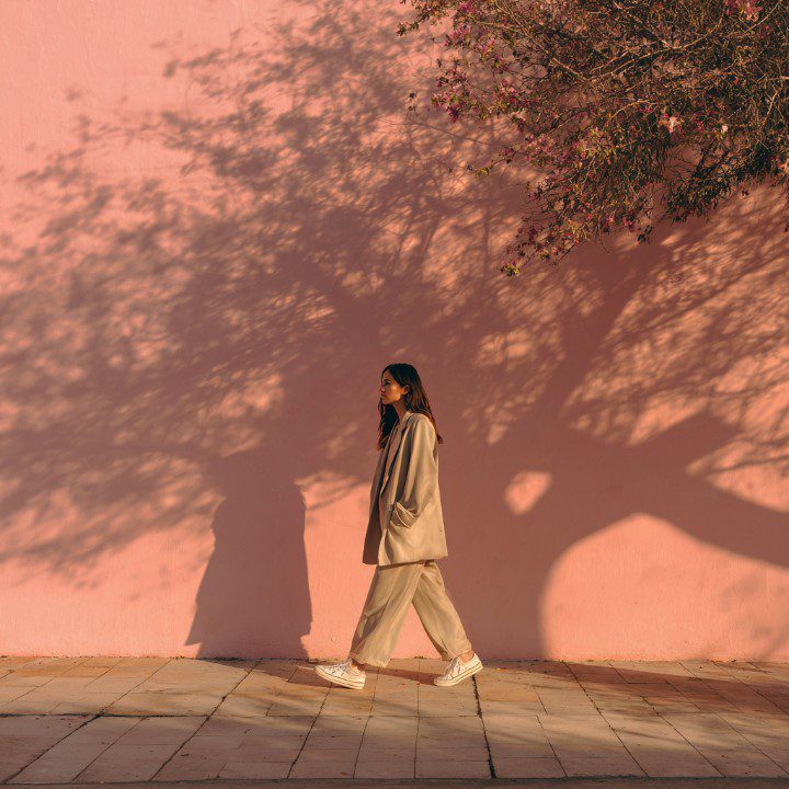 Stylish woman in beige outfit walking along a pink wall under warm sunlight with artistic tree shadows, symbolizing elegance and minimalism.