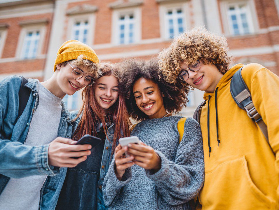 Group of diverse teenage students standing outside, smiling and using smartphones, showcasing youth, technology, and friendship.