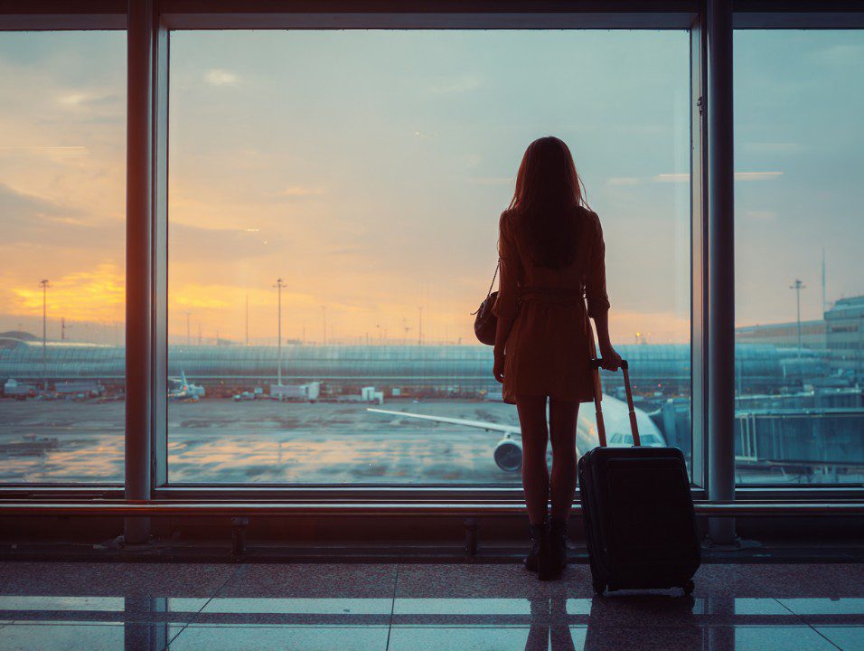 Silhouette of a woman standing with luggage at an airport window at sunrise, waiting for her flight.