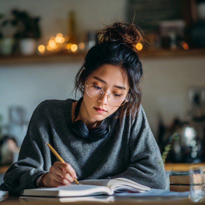 Young woman with glasses and headphones writing in a notebook at home, representing focused study and remote learning in a cozy setting.