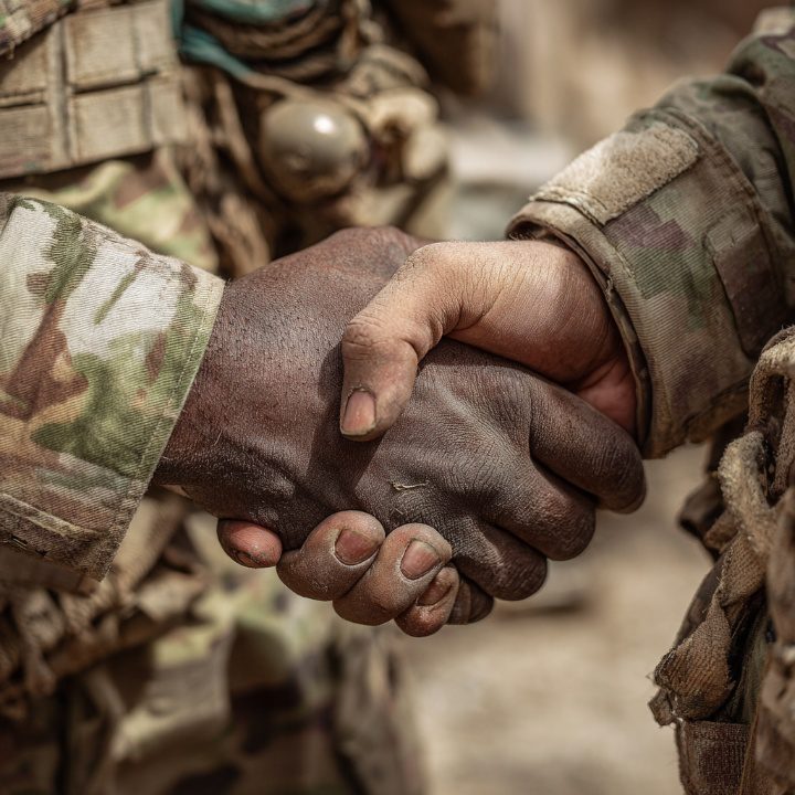 Close-up of two soldiers from different backgrounds shaking hands while wearing camouflage military uniforms, symbolizing unity and respect.