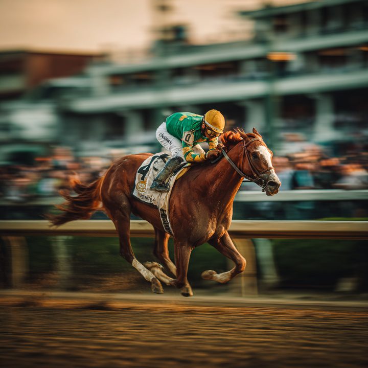 Side profile of a thoroughbred racehorse in full gallop with a jockey in green and yellow silks, captured with motion blur on a racetrack during golden hour.