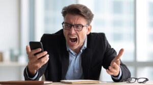 Frustrated businessman in glasses shouting at a smartphone while sitting at an office desk.