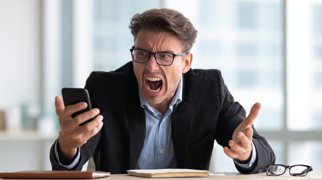 Frustrated businessman in glasses shouting at a smartphone while sitting at an office desk.